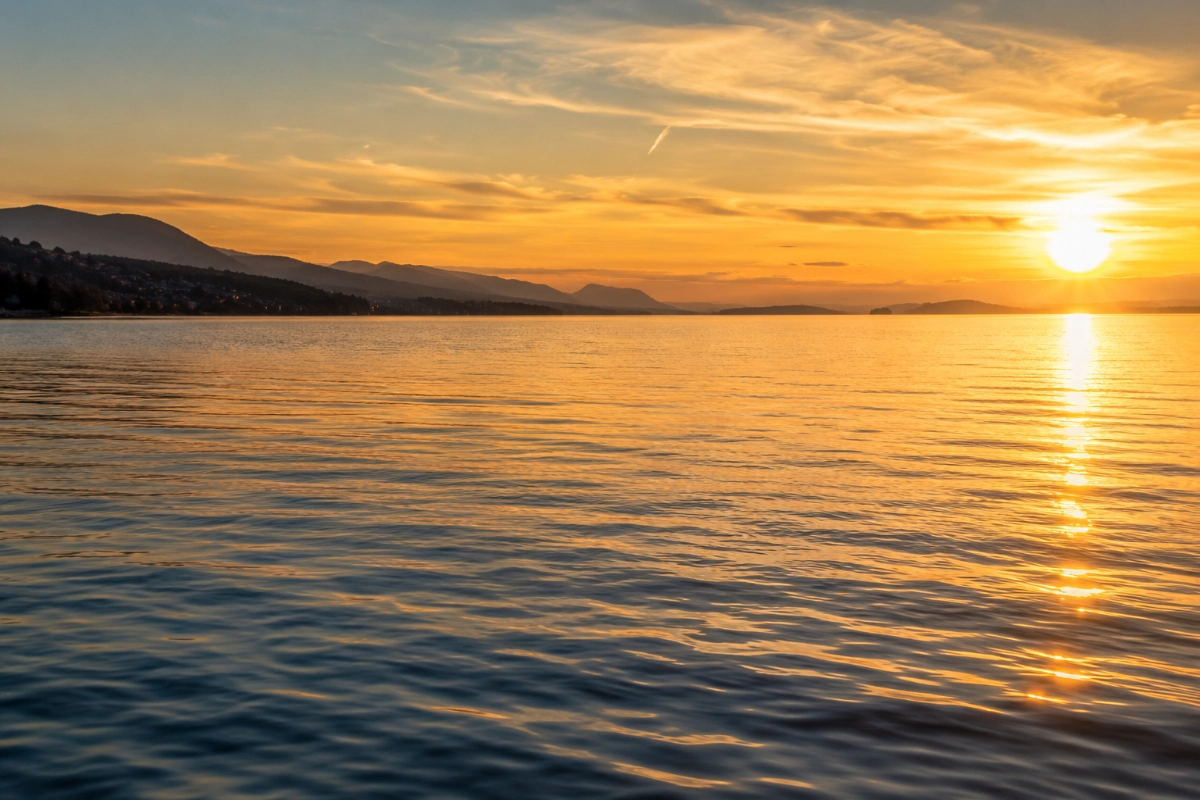 Open water under golden evening light, gentle ripples reflecting the sky, distant shoreline fading into haze, eye-level perspective, atmosphere of hope and possibility.