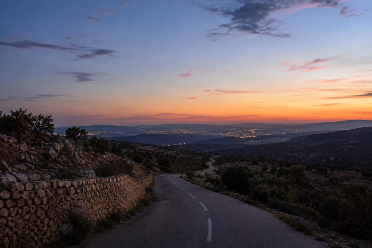Quiet country road at dusk beneath a fading sky, distant lights appearing on the horizon, wide perspective, atmosphere of longing and pilgrimage — Photorealistic, authentic editorial photography, natural colors, realistic lighting, documentary style, premium magazine photography, no painting, no illustration, no digital art, no fine art composition, no symbolic artwork, no text, no watermark.